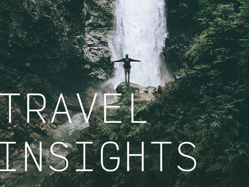 A person at the base of a waterfall with their arms stretched out wide and the caption "Travel Insights".