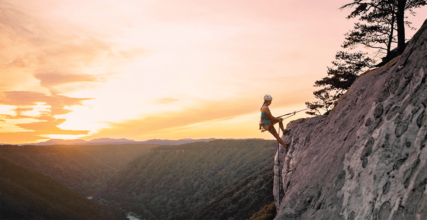 A woman rock climbing in West Virginia with a sunset in the background.